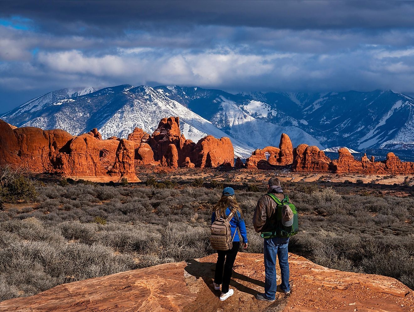 Couple enjoying beautiful mountain view on hiking trip in Utah. The Windows Section of the park, snow covered La Sal Mountains in the background. Arches National Park ,Moab, Utah.
