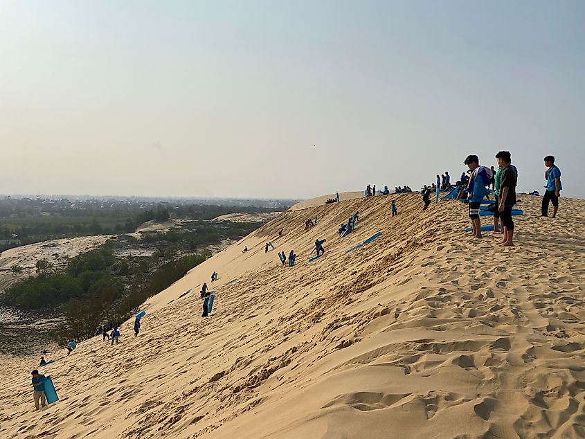 A group of school children take turns sledding down a large set of sand dunes.